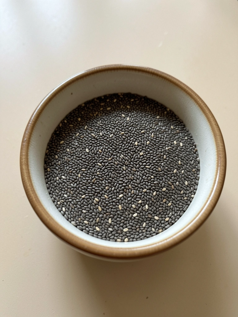 Pequenos grãos de chia escuros e brilhantes acumulados em uma tigela de cerâmica clara.
